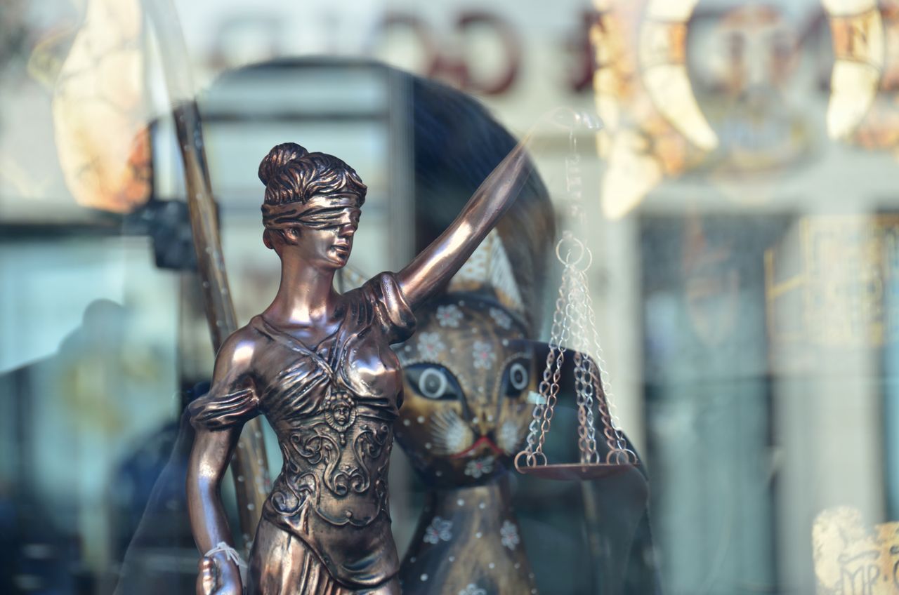 A close-up of a Lady Justice figurine with a blurred background, captured through a reflective window in Poland.